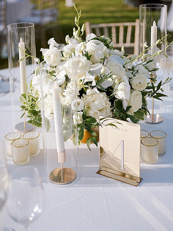 Wedding centerpiece with a white rose centerpiece, greenery and white taper candles in glass hurricanes on a white tablecloth at an outdoor lawn reception