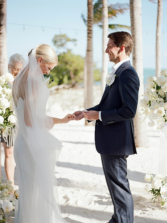 Wedding vows as bride and groom hold hands, long bridal veil flowing during an outdoor wedding ceremony on sand with palms and ocean backdrop