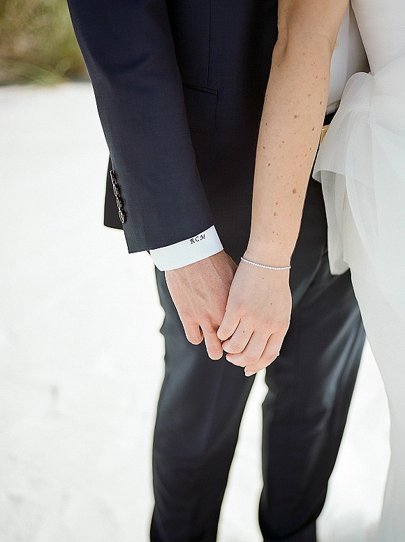 Couple holding hands, wedding handholding photo showing bride’s white dress and bracelet beside groom’s suit cuff on a green outdoor walkway