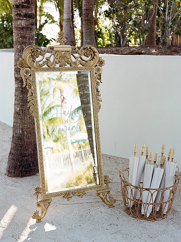 Wedding welcome sign on an ornate gold mirror frame with calligraphy lettering beside parasols in a wicker basket, set by palm trees and a white stucco wall