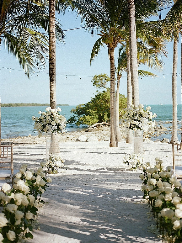 Beach ceremony setup with oceanfront wedding ceremony aisle lined with white rose markers, bistro chairs, and string lights by palm trees