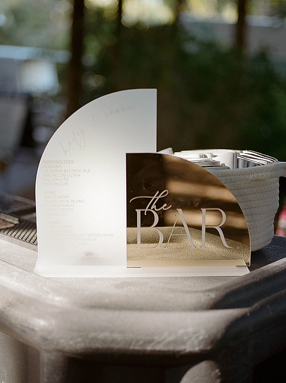 Wedding bar sign on a clear acrylic bar sign beside a drink menu card, bar tools, and tray in window light with greenery behind