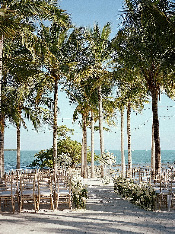 Beach ceremony setup with oceanfront wedding ceremony seating, white rose aisle flowers and string lights on sand with palms and shoreline backdrop