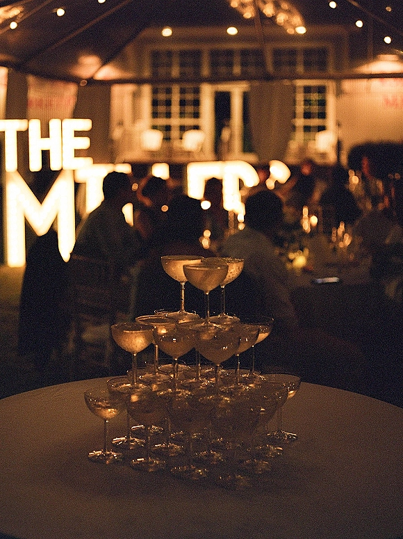 Champagne tower of stacked coupe glasses on a round table with string light accents under a tent canopy at a night reception