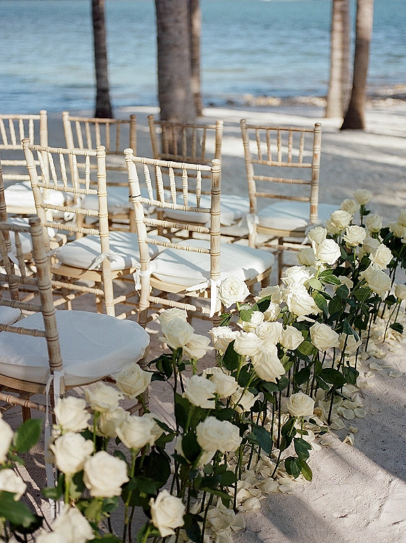 Ceremony aisle decor with white rose arrangements and rose petals lining a sandy beach aisle, chiavari chairs facing the oceanfront