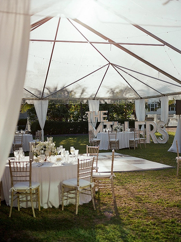 Outdoor wedding reception under a clear top wedding tent with string lights, round tables in white linens, gold Chiavari chairs, and dance floor on a lawn