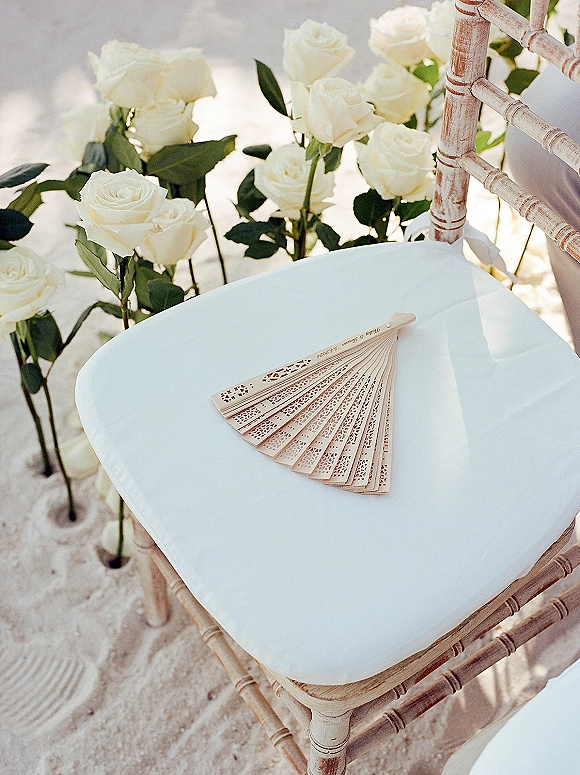 Wedding ceremony fans resting on chiavari chairs with wooden hand fan, white roses and greenery accents on sandy outdoor aisle in sunlight