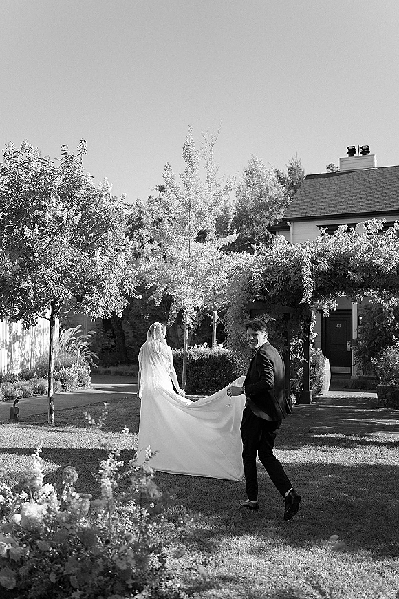 Couple portrait in a black and white wedding photo, bride and groom walking away on a garden lawn as he lifts her long veil train