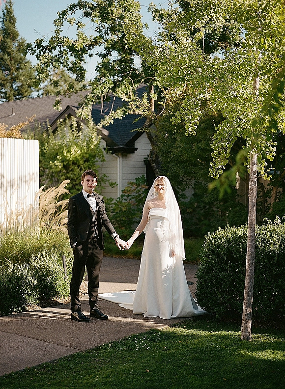 Couple portrait of bride and groom holding hands, her long veil and dress train flowing on a garden path with trees and shrubs behind