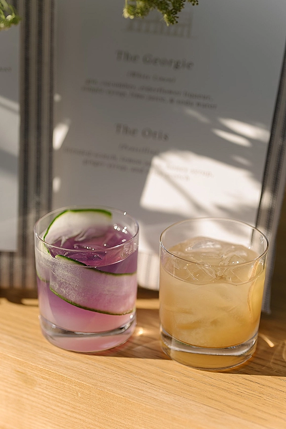 Signature cocktails beside a wedding signature drinks menu card, with ice-filled glasses and cucumber ribbon garnish on a sunlit wood tabletop