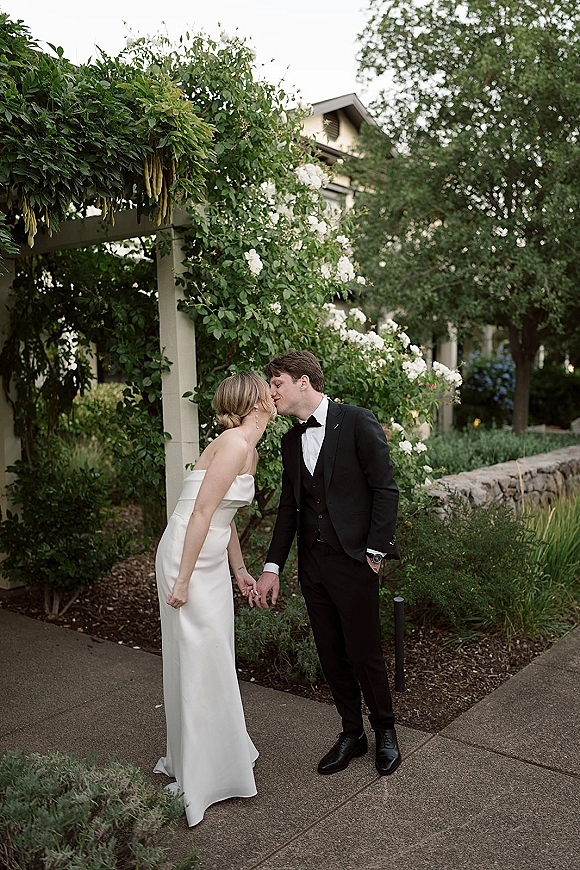 Wedding kiss portrait of bride and groom kissing, holding hands under a white rose pergola, with strapless dress and black tuxedo bow tie