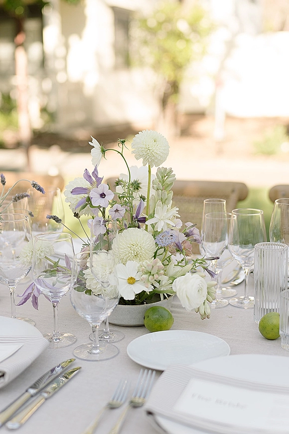 Reception tablescape with outdoor wedding table setting, pastel floral centerpiece in ribbed vase, linen cloth, glassware, menus, and limes on sunlit patio