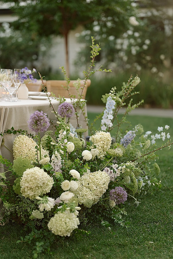 Reception tablescape with hydrangea and allium centerpieces, linen tablecloth, glassware, and rattan chairs on a garden lawn
