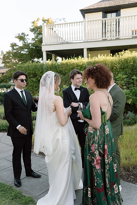 Wedding cocktail hour with wedding guests mingling as the bride in a lace veil chats near garden hedges, guests holding champagne flutes