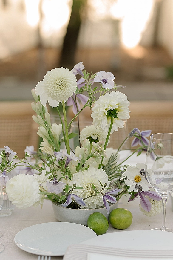 Reception centerpiece with white and lilac flowers in a low bowl vase on a linen table, with votive candle, wine glass, and limes on an outdoor patio