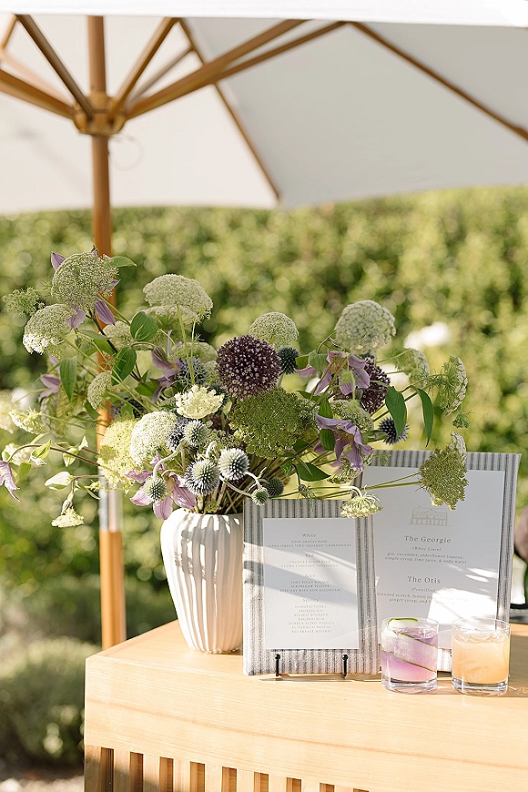 Reception drink station with wedding cocktail menu on an acrylic stand, rocks glass cocktails, and wildflower vase under a bar umbrella outdoors