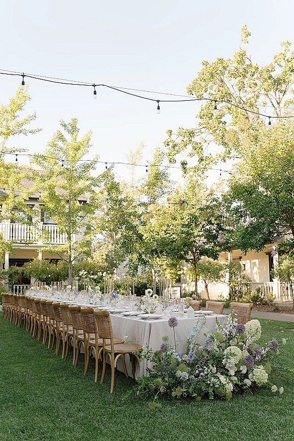 Outdoor reception tablescape with a long banquet table wedding setup, wicker chairs, candles and floral centerpieces under string lights on a garden lawn