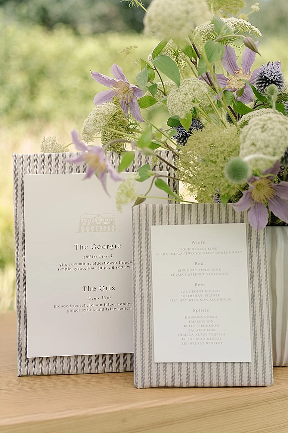 Wedding bar menu with signature cocktail options on striped linen boards, framed by clematis and thistle flowers on a wooden table outdoors