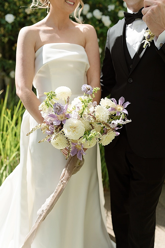 Wedding couple portrait of bride holding a white and lavender bouquet with trailing ribbon beside groom in black tuxedo amid sunlit garden greenery