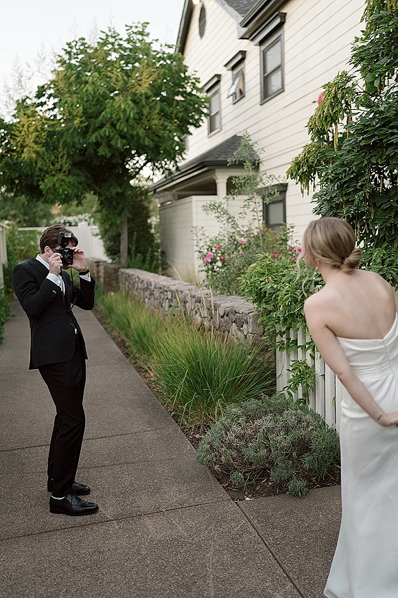 Wedding photographer with flash attachment shooting a groom in black tux and bride in strapless gown by a white picket fence walkway