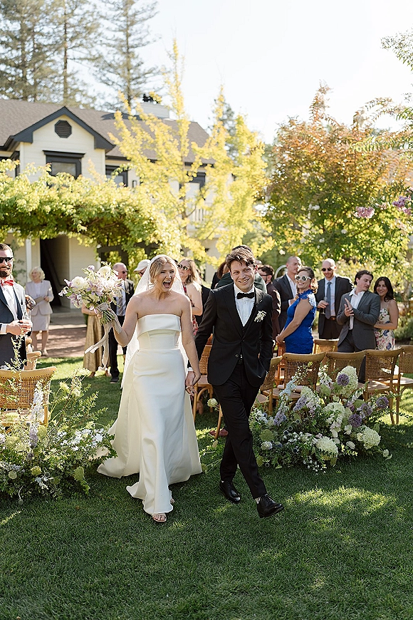 Wedding recessional as bride and groom walk the aisle holding hands, bouquet in hand, guests cheering on a garden lawn with trees and house backdrop