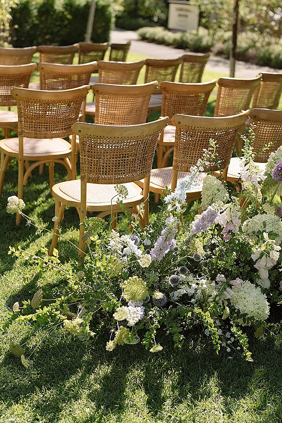 Ceremony seating with rattan wedding chairs in neat rows on a grassy lawn, framed by low white and lavender aisle florals and greenery