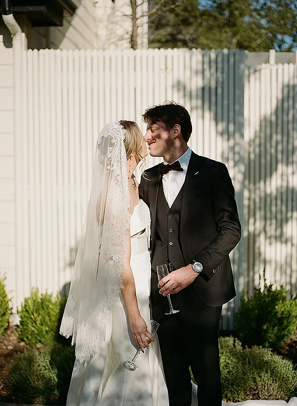 Couple portrait of bride and groom holding champagne flutes, her lace veil flowing as they smile by a white picket fence outdoors