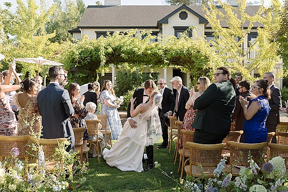 Wedding recessional as newlyweds share a just married kiss in a garden aisle, veil flowing past wicker chairs and applauding guests under pergola
