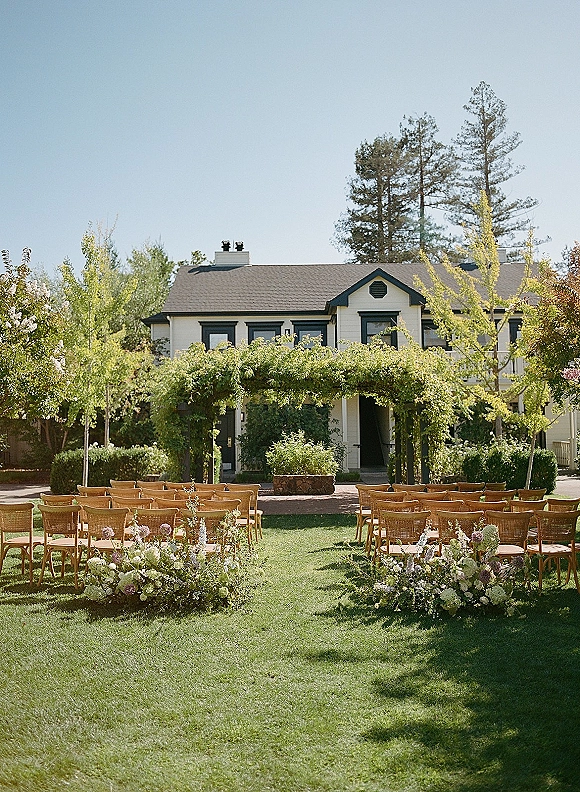 Ceremony setup with outdoor wedding ceremony seating, wood chairs flanking a floral aisle and greenery arch on a garden lawn by a house facade