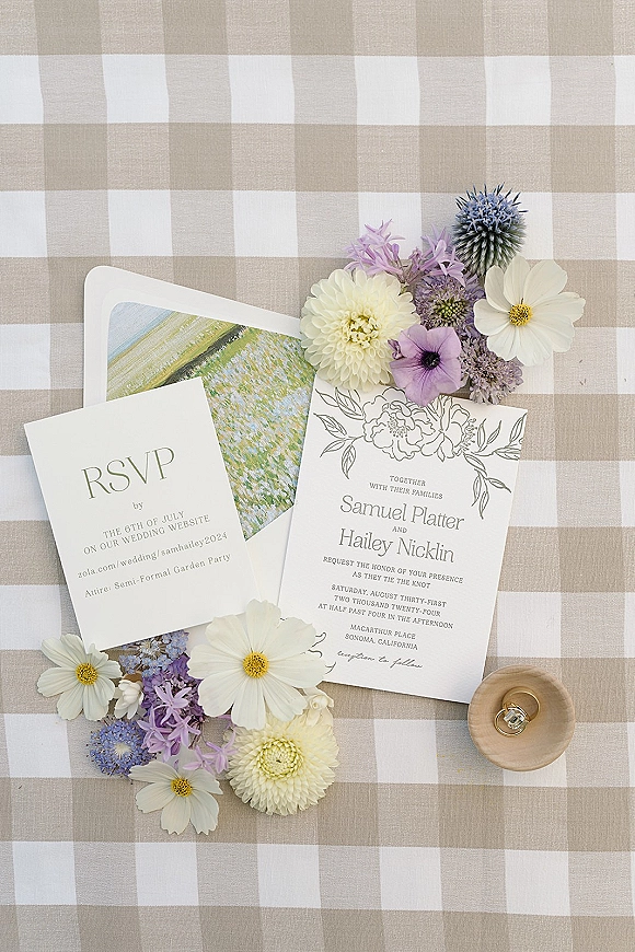 Invitation suite wedding invitation flat lay with floral line art, RSVP card, envelope, and engagement ring on neutral gingham tablecloth