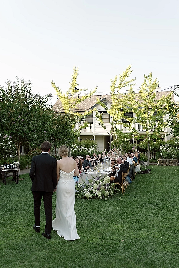 Reception entrance as bride and groom walk past a long banquet table with floral centerpieces and string lights on a garden lawn