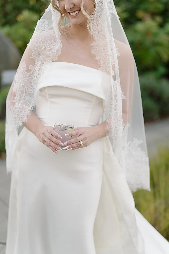 Bridal portrait of a smiling bride in a strapless wedding dress with lace veil, holding a cocktail glass on an outdoor walkway with greenery