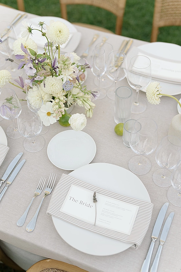 Reception tablescape with neutral linen, striped napkins, place cards and menus, lavender sprigs, and white-green flowers on a green lawn
