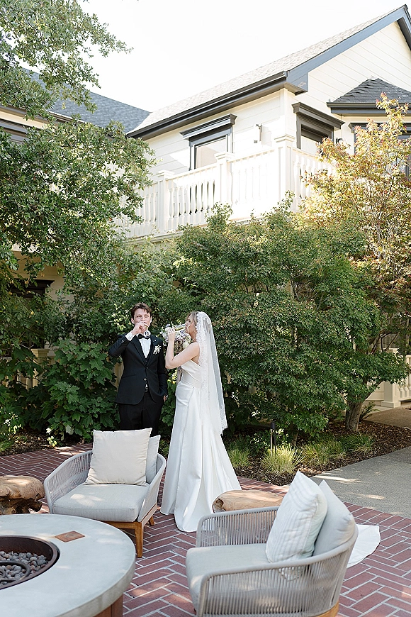Couple toast with champagne flutes, bride in long veil and satin wedding dress beside groom in black tuxedo on a brick patio lounge by fire pit