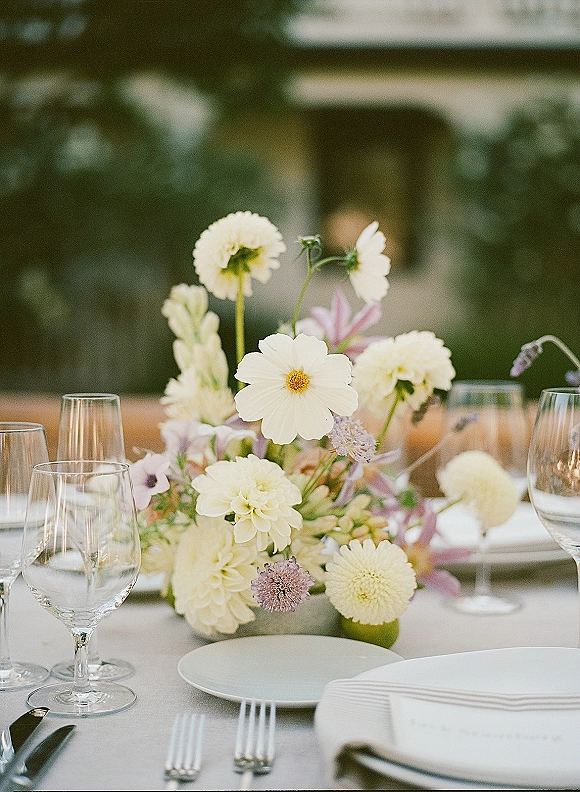 Reception tablescape with wedding table centerpiece of low white, pale yellow, and lavender florals, bud vases, and clear stemware on a patio table