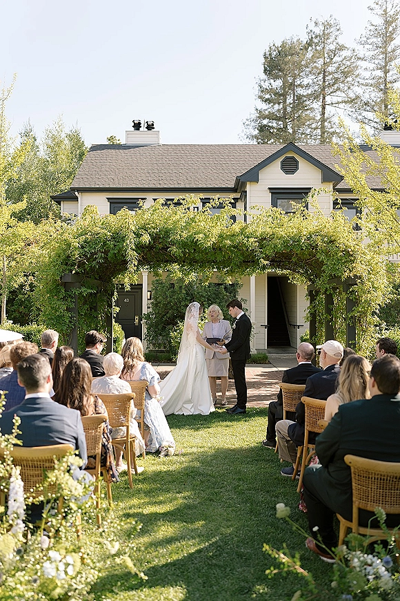 Wedding ceremony with bride in long veil and groom in tuxedo, guests seated by aisle floral arrangements under a greenery arbor on a garden lawn