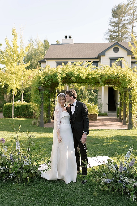 Couple portrait of bride and groom kiss, holding champagne flutes beneath a greenery-covered pergola on a sunny garden lawn with floral arrangements