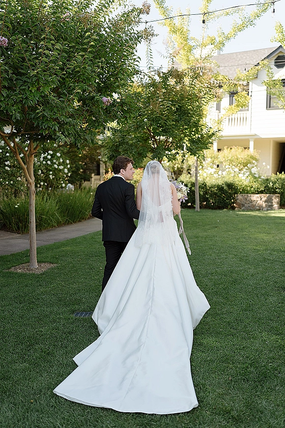 Couple portrait of bride and groom walking away on a garden lawn, her long veil and train flowing as he holds her under string lights