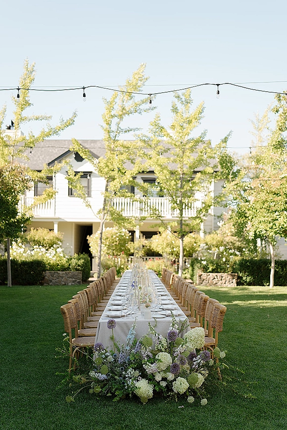 Reception tablescape with a long banquet table wedding set in white linen, rattan chairs, bud vases, greenery garland, and string lights on a lawn