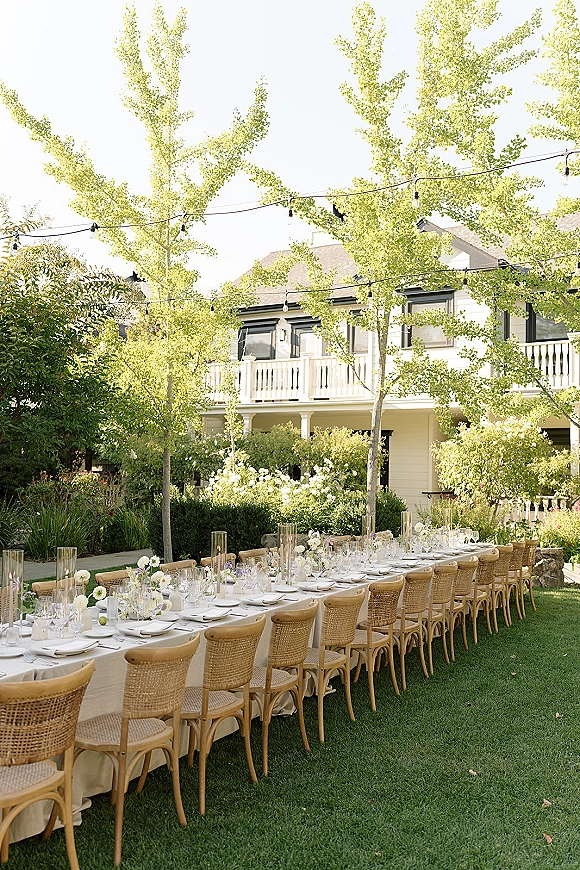 Outdoor reception tablescape with a long banquet table wedding setup, beige linens, rattan chairs, white florals and string lights on a garden lawn