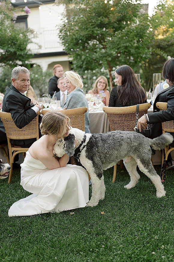 Bride with dog cuddling close in a strapless wedding dress at an outdoor reception, with rattan chairs, dinner tables, and string lights behind
