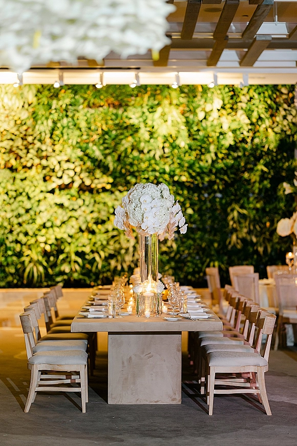 Reception tablescape with a long banquet table setup, tall white floral centerpieces, candlelit place settings, and a greenery wall backdrop indoors