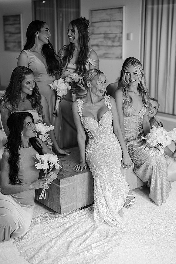 Bride with bridesmaids laughing in a hotel suite, wearing a lace wedding dress and holding bouquets by a window with curtains