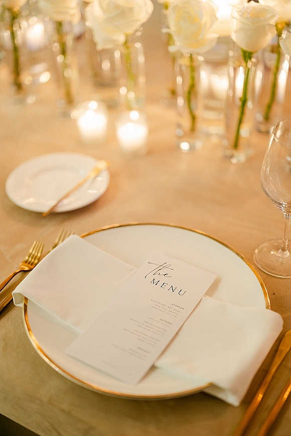 Reception tablescape with wedding place setting, gold-rim plates, menu card on napkin, gold flatware, white roses, and candlelight on neutral linen