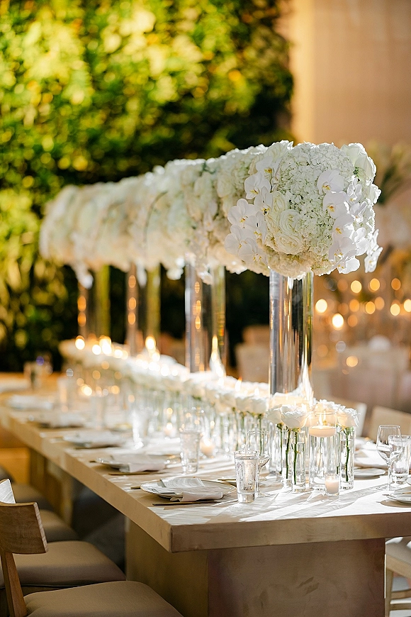 Reception tablescape with white wedding tablescape tall glass vases of white florals, floating and votive candles, set on an outdoor patio by a hedge wall