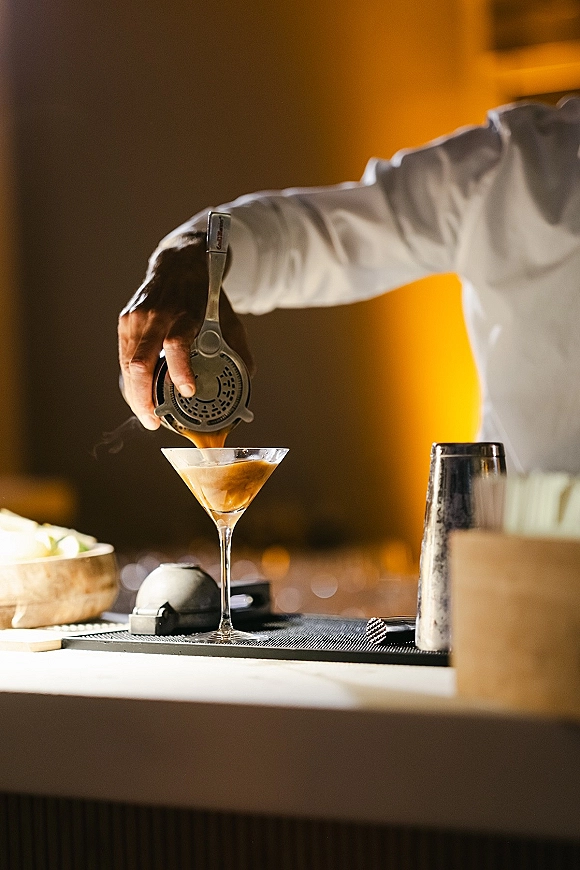 Wedding cocktail bar with a cocktail coupe glass, strainer, and shaker tin arranged on a bar mat under warm indoor lighting