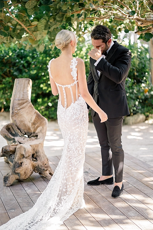 First look moment as bride in a lace open-back gown walks up behind groom in tuxedo wiping tears on a wood deck under trees