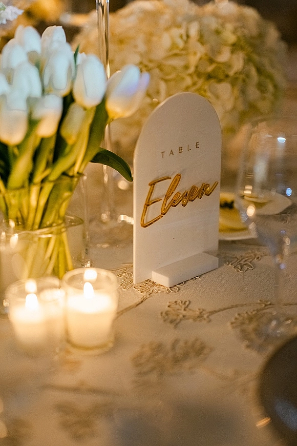 Wedding table number acrylic table number sign with gold lettering beside tulips and hydrangeas, votive candles, and goblets in warm light