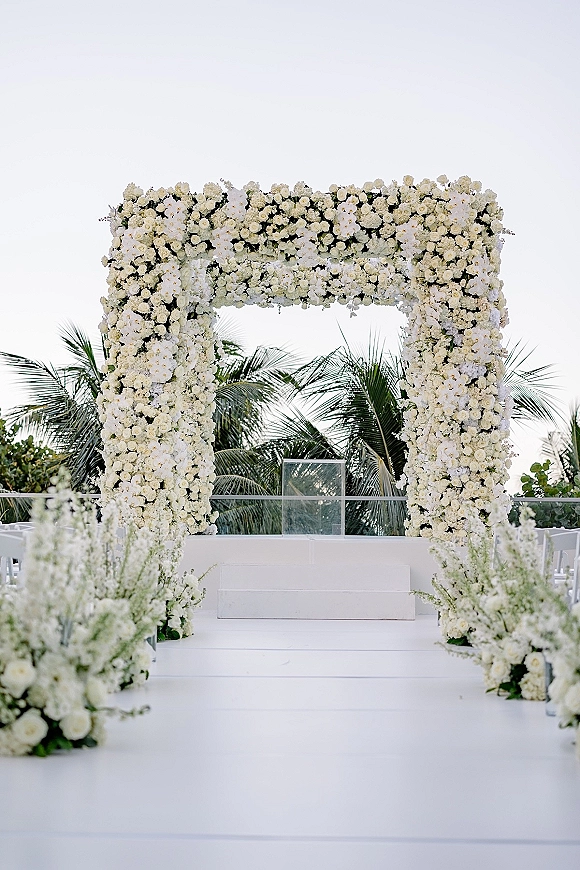 Wedding ceremony arch with a white floral wedding arch of roses and orchids, greenery, and aisle florals on a terrace with palm trees and sky