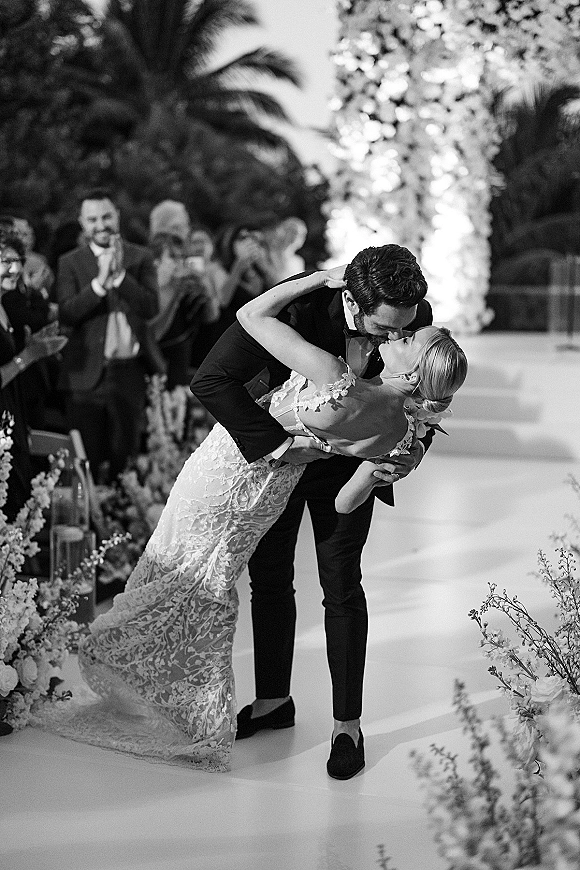 Wedding kiss portrait as the groom dips the bride in a lace off-the-shoulder gown, tuxedo bow tie, palm-lined aisle with guests behind